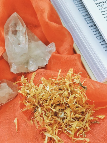 Dried Calendula flowers on an orange scarf with clear quartz and an open herbalism book